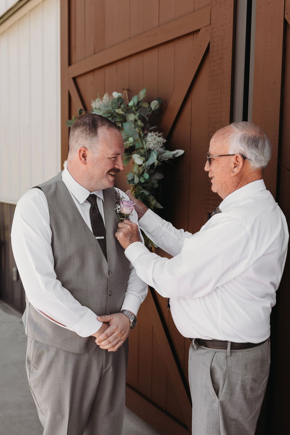 An older man pins a boutonniere on a groom in a gray vest and black tie outside a rustic barn wedding venue.