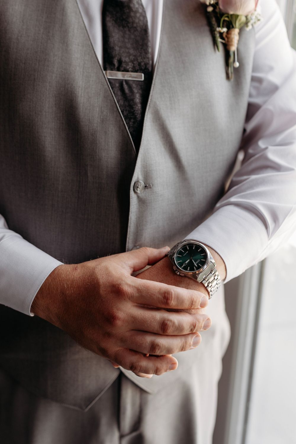 Close-up of groom in gray vest adjusting sleeves wearing elegant silver watch with dark blue face.