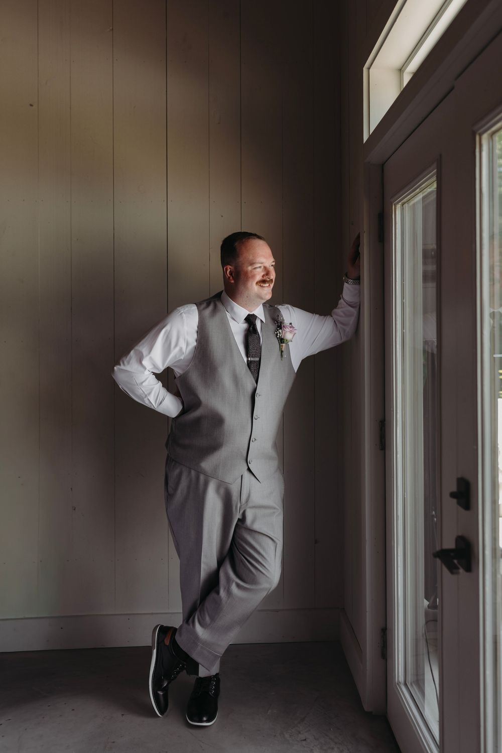 Smiling groom in gray vest and tie with boutonniere leans against wall by glass door in natural light.