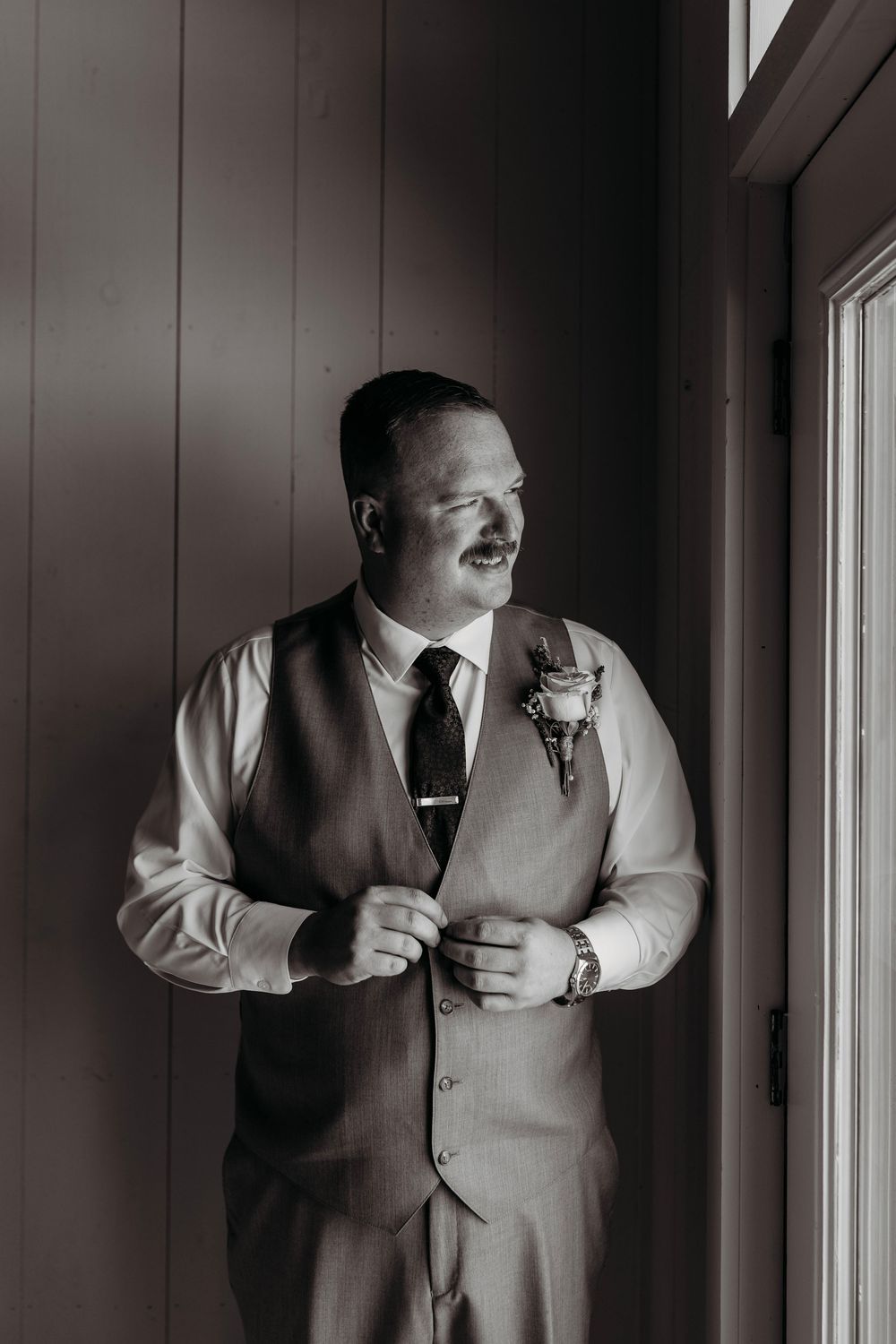 Groom in vest and tie adjusting buttons by window in black and white wedding portrait with boutonniere.