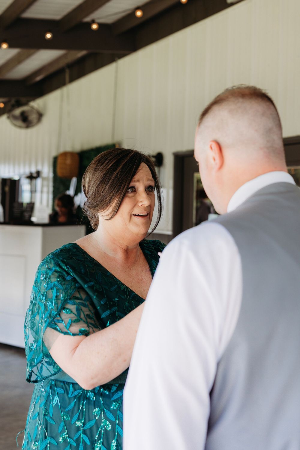 Woman in teal lace dress talking with groom in gray suit at wedding venue.