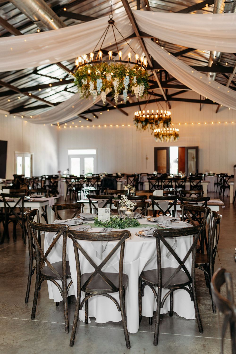 Elegant barn wedding reception with floral chandeliers, string lights, draped fabric ceiling, and rustic cross-back chairs at round tables.