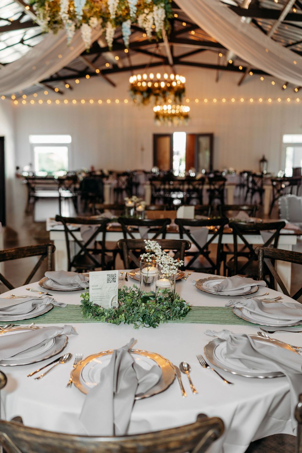 Elegant wedding reception hall with round tables set with gray linens, greenery centerpieces, candles, and string lights overhead.