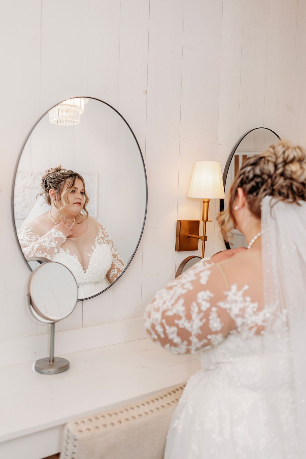 Bride in lace wedding gown adjusts jewelry while gazing at her reflection in an oval vanity mirror.