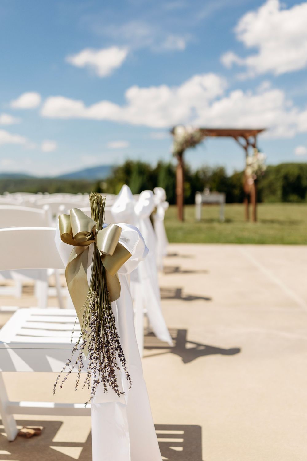 White wedding chairs with gold ribbon and lavender bouquet decor line an outdoor aisle under a sunny blue sky.