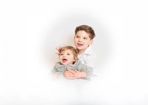 Two young siblings smile joyfully, older boy hugging toddler, in a bright white studio portrait.