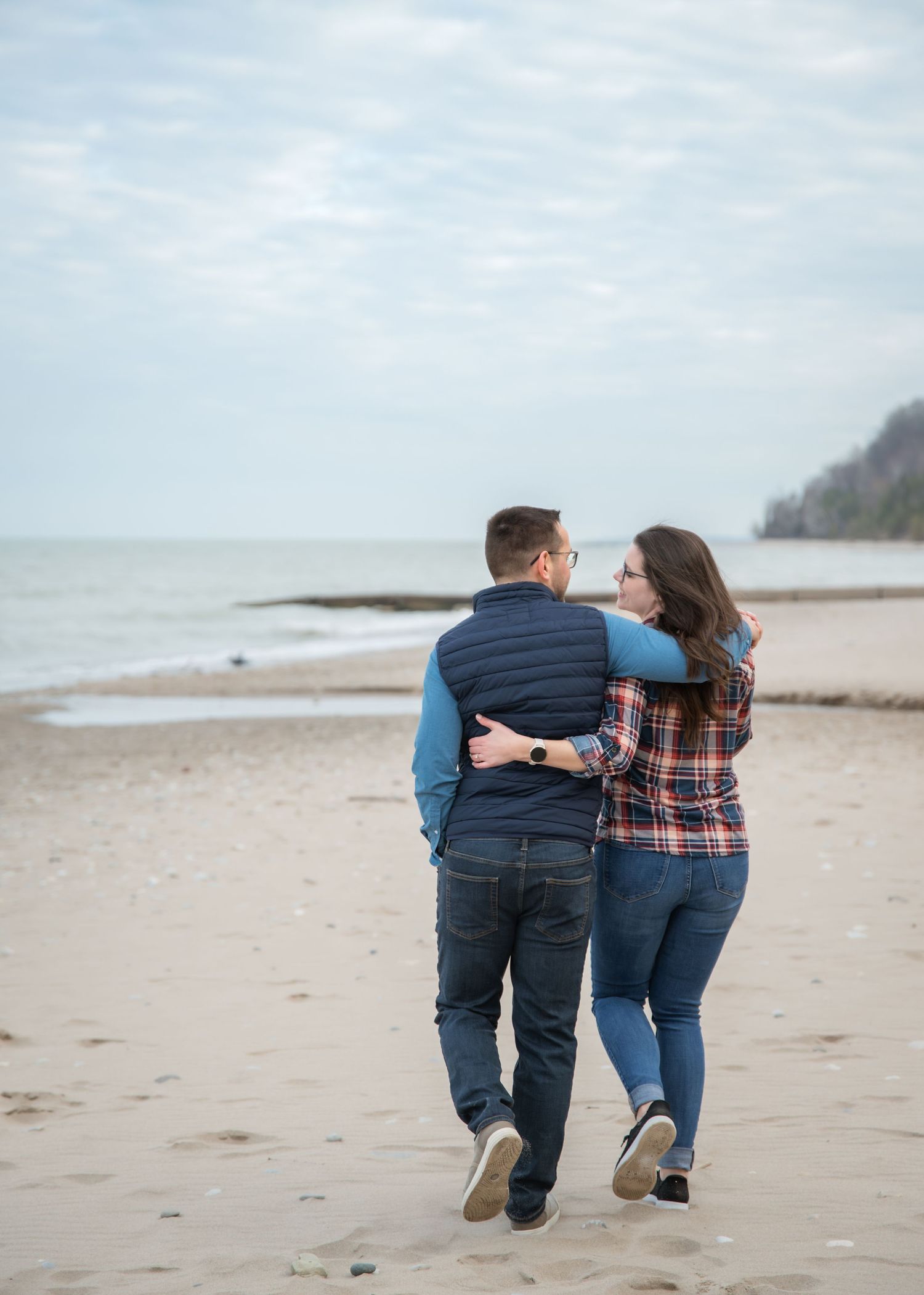 Couple walking arm in arm on a sandy beach, wearing casual fall attire, with calm ocean waves in the background.