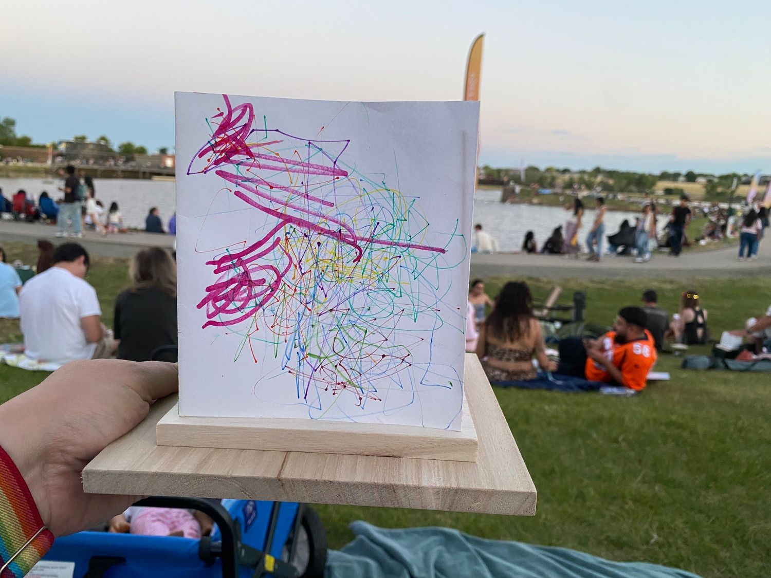 Hand holding a child's colorful drawing of a tree with clouds and sun on a wooden easel at an outdoor park gathering.