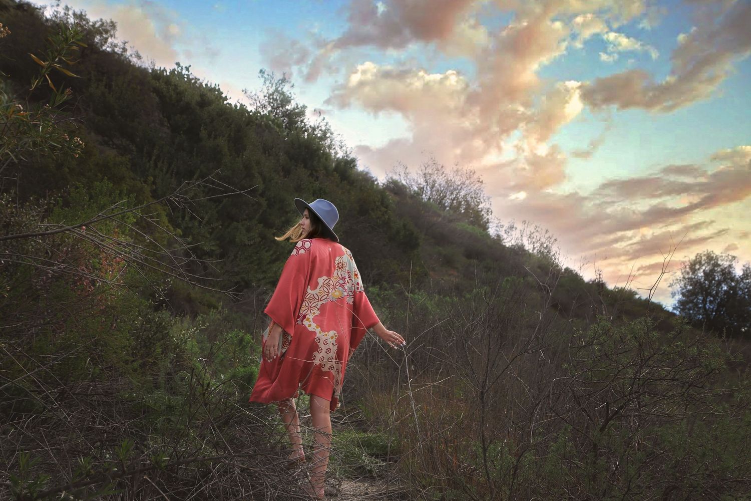 Woman in a red floral kimono and wide-brim hat walks up a grassy hillside under a colorful cloud-filled sky.