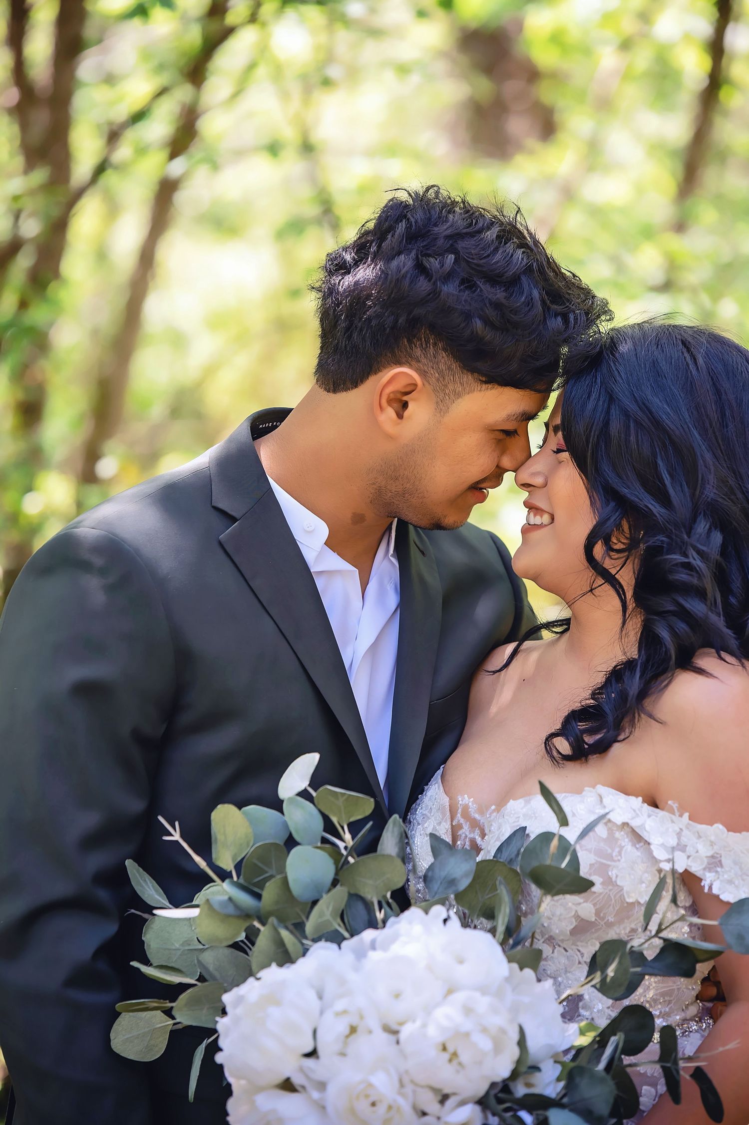 Newlyweds share tender moment with white floral bouquet among green foliage.