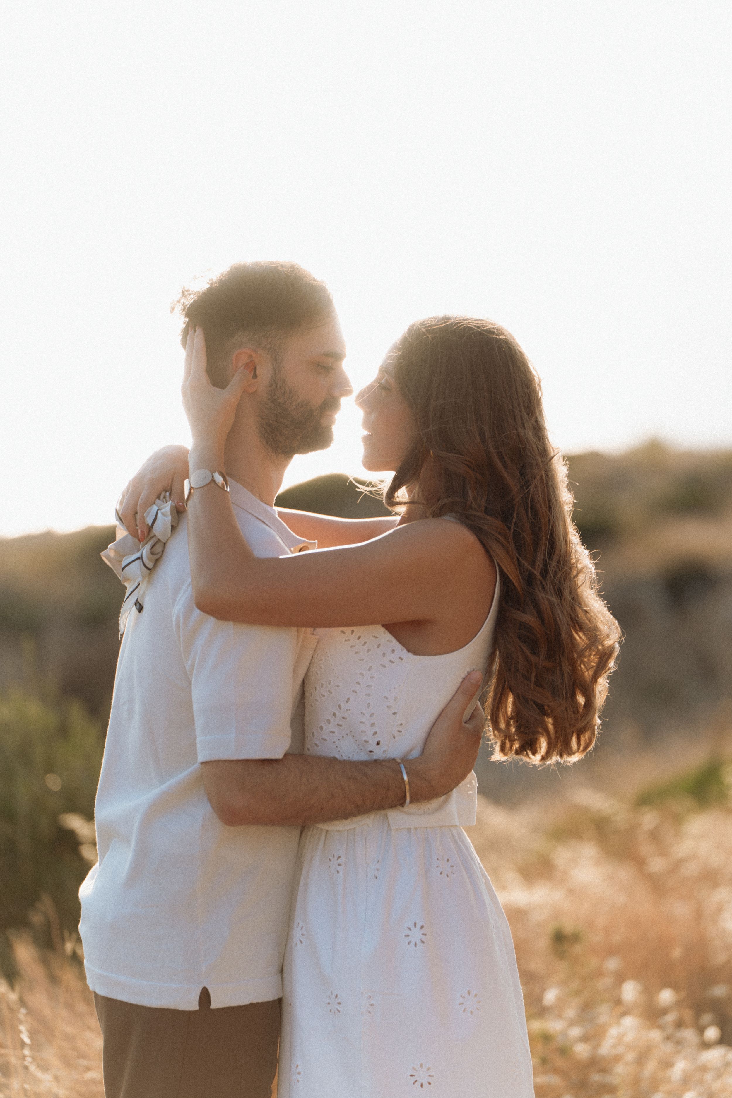 Romantic couple embracing in golden sunlight, wearing white outfits in a scenic outdoor field.