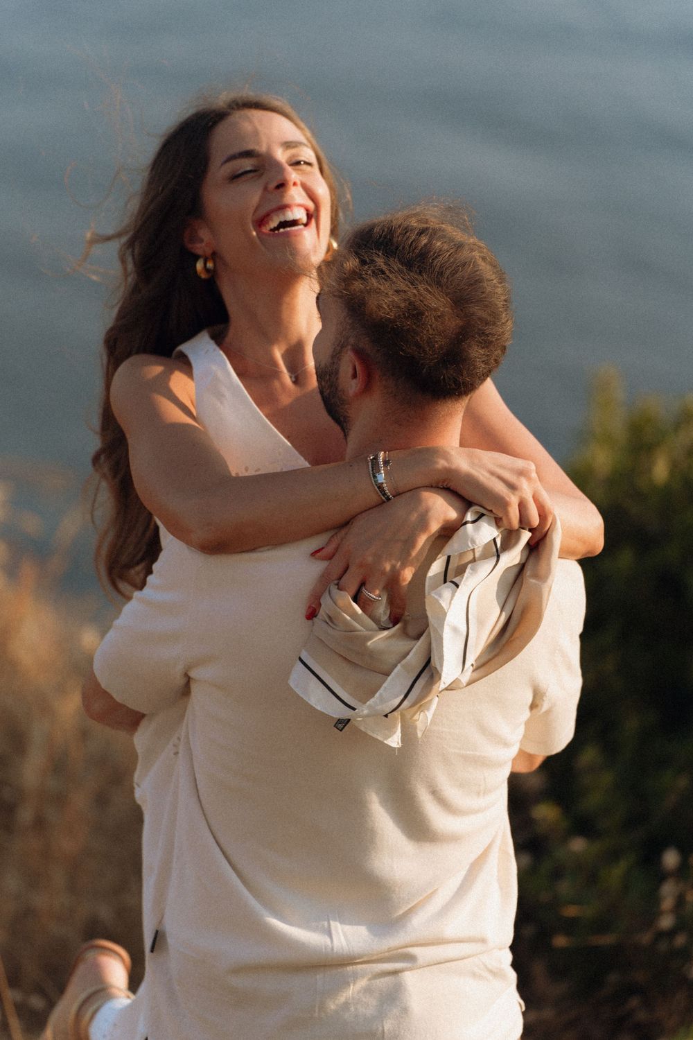 A joyful couple embraces and laughs together outdoors near a scenic waterfront at golden hour.
