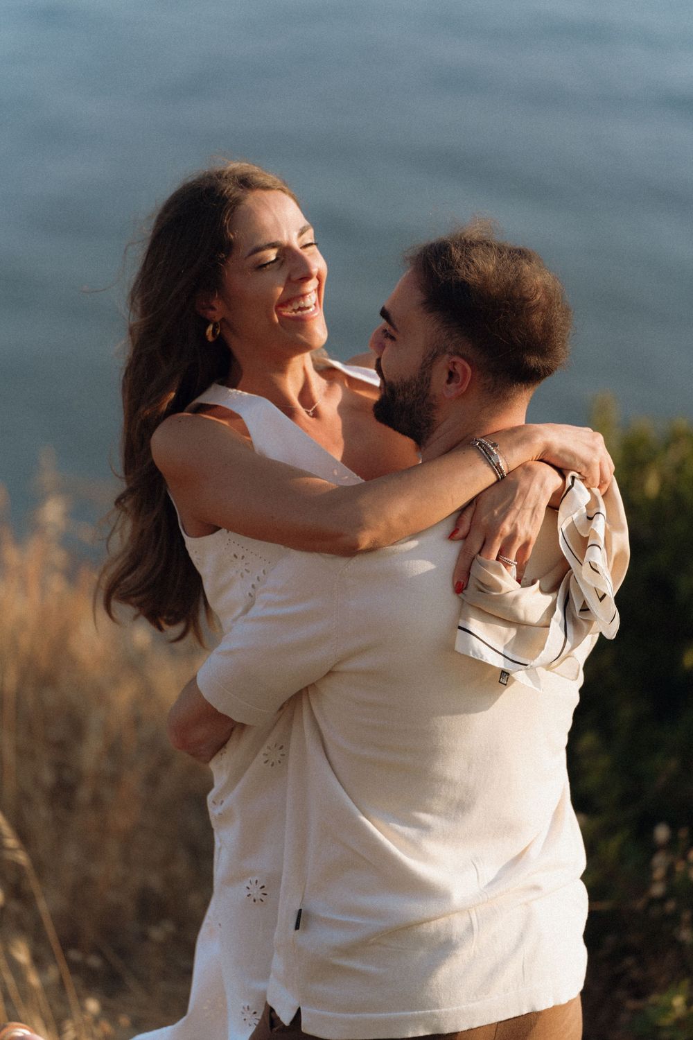 A joyful couple embraces and laughs together on a coastal cliff at golden hour, with the ocean in the background.