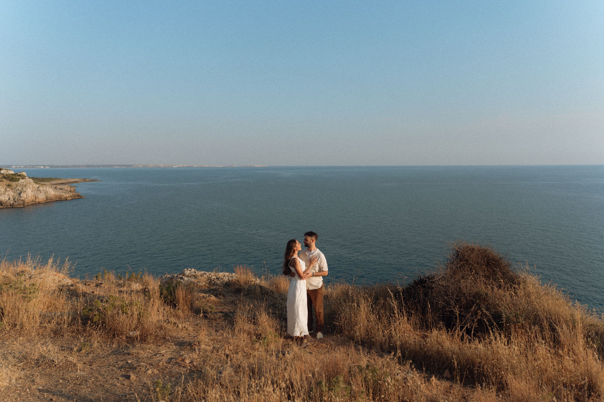 Couple embracing on a scenic coastal cliff with golden grass, overlooking a calm blue sea under a clear sky.