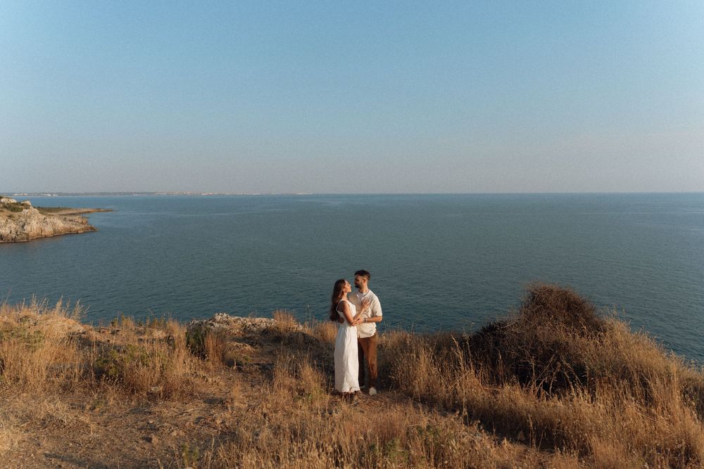 Couple embracing on a scenic coastal cliff with golden grass, overlooking a calm blue sea under a clear sky.