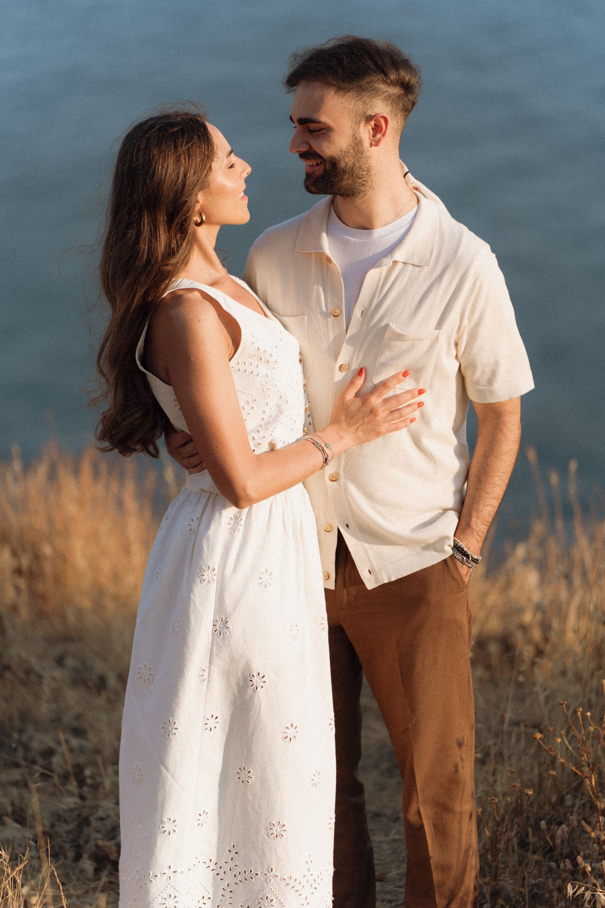 Couple in love standing outdoors near water, woman in white eyelet dress, man in cream shirt and brown pants.