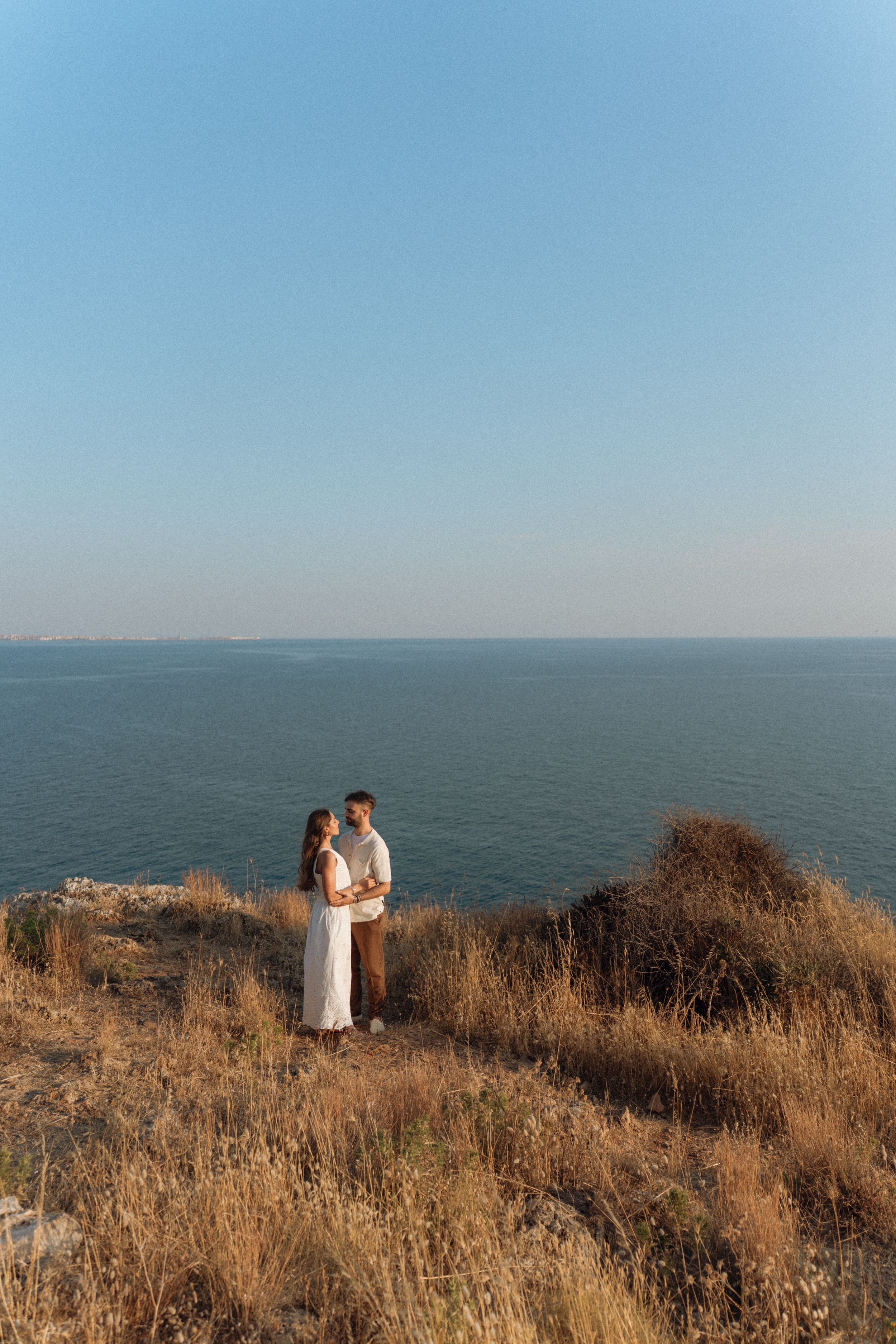 Couple embracing on a cliff overlooking the ocean at sunset, surrounded by golden grass under a clear blue sky.
