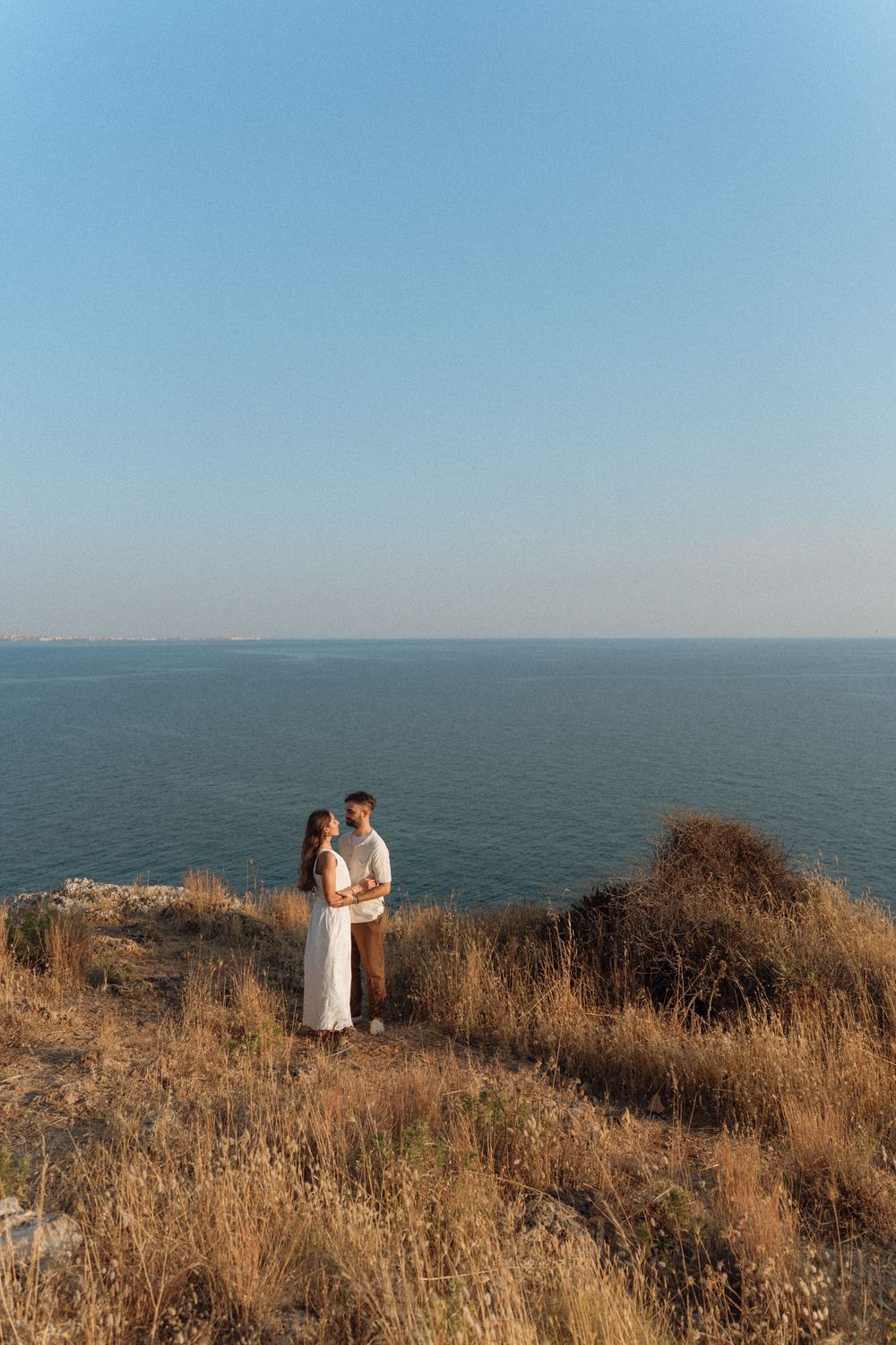 Couple embracing on a cliff overlooking the ocean at sunset, surrounded by golden grass under a clear blue sky.