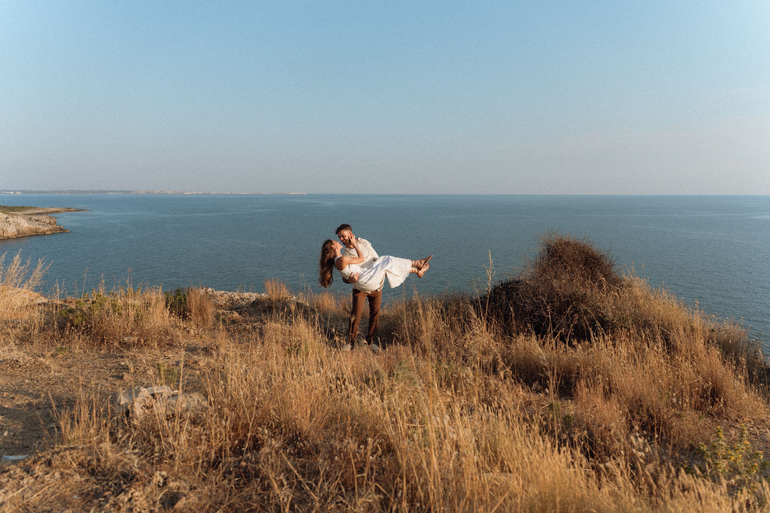 Couple embracing on a scenic coastal cliff with golden grass, blue sea and clear sky in the background.