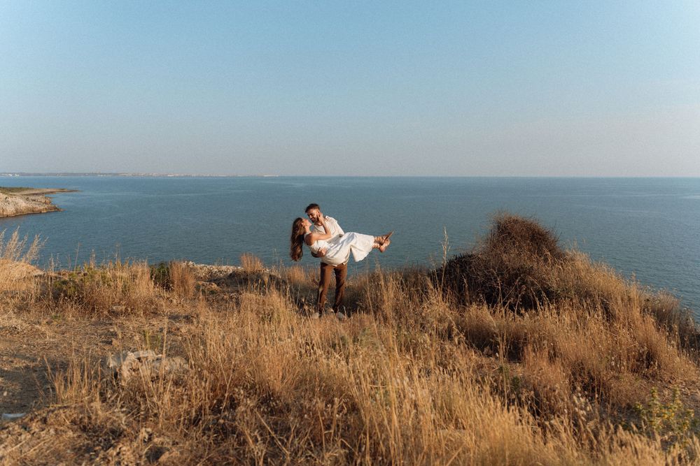 Couple embracing on a scenic coastal cliff with golden grass, blue sea and clear sky in the background.