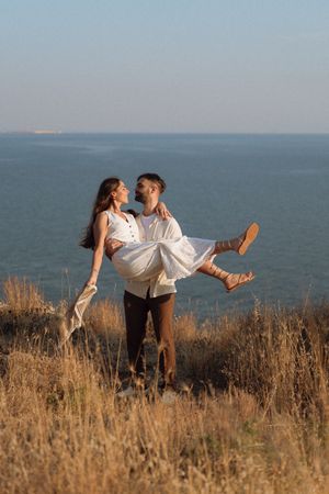 A man carries a woman in his arms on a coastal hillside, both dressed in white, with the ocean behind them.