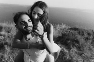 Black and white photo of a smiling man giving a woman a piggyback ride by the ocean.