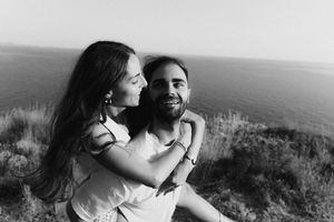 Black and white photo of a happy couple embracing outdoors on a cliff overlooking the ocean.