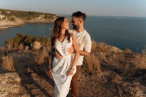 A romantic couple embraces on a rocky coastal cliff overlooking a stunning blue ocean at sunset.