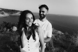 Black and white photo of a smiling couple standing on a coastal cliff with ocean views in the background.