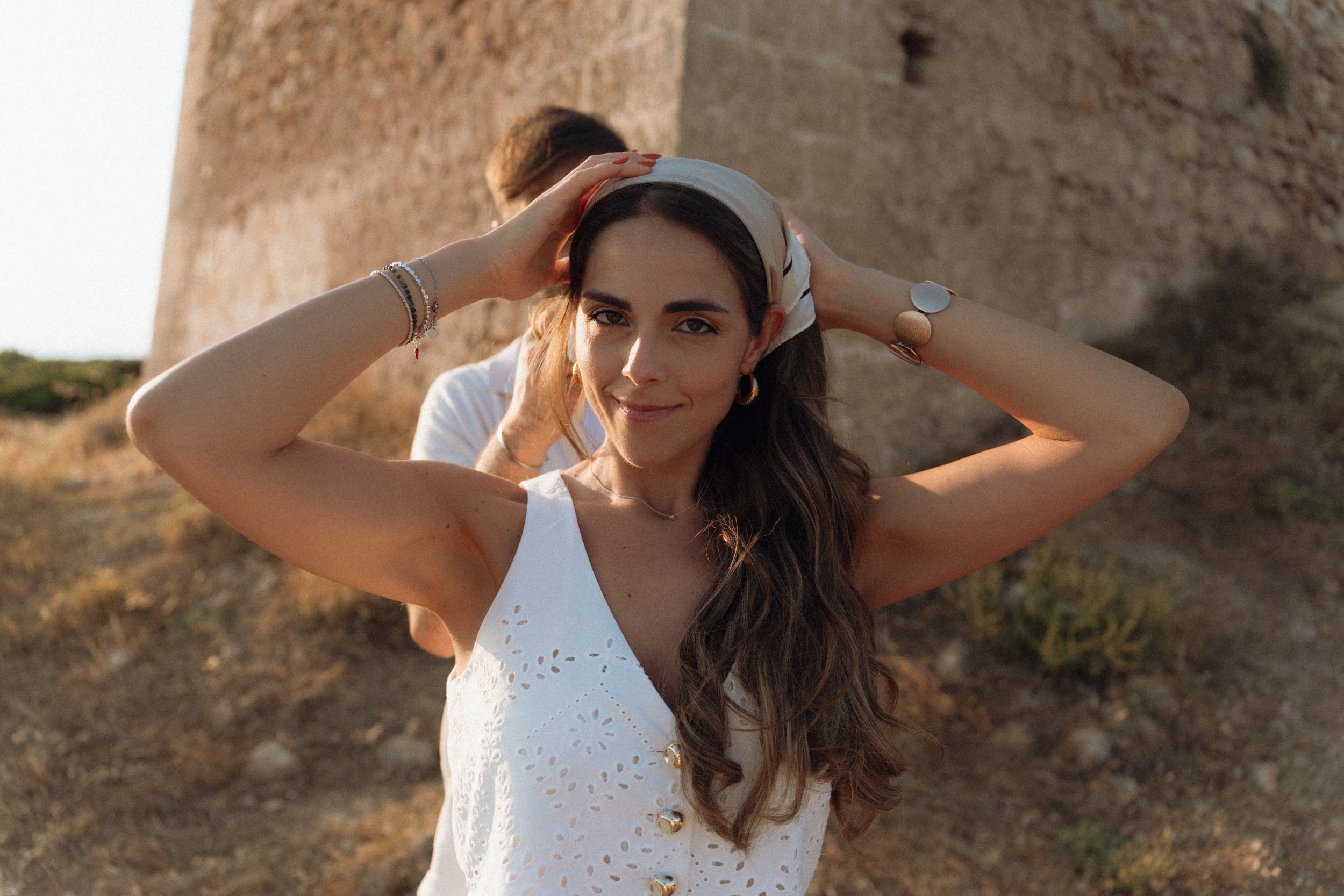Woman in white dress adjusting headband outdoors near ancient stone tower in warm sunlight.
