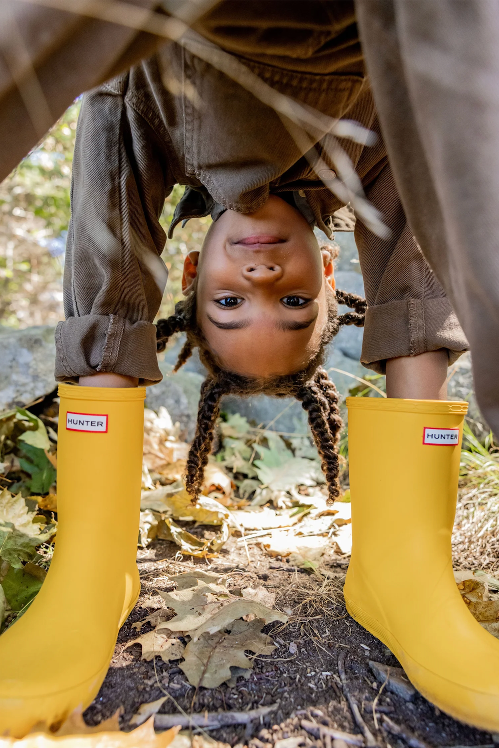 Child bending over in yellow Hunter rain boots with braided hair hanging upside down outdoors.