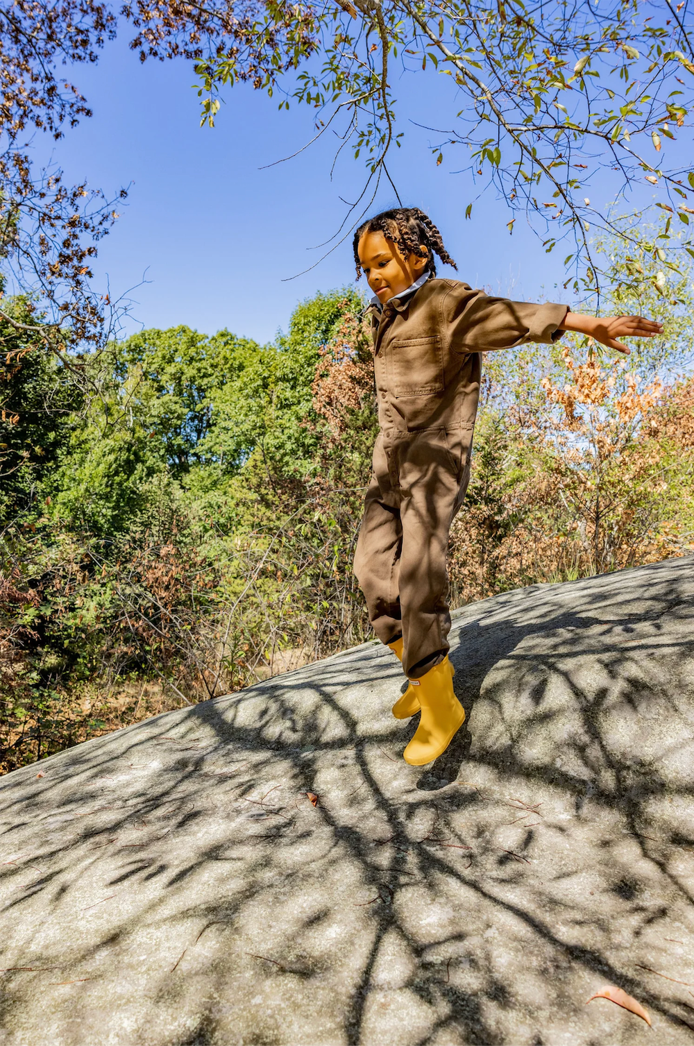 Child in yellow Hunter boots balances on a large rock surrounded by colorful autumn foliage.