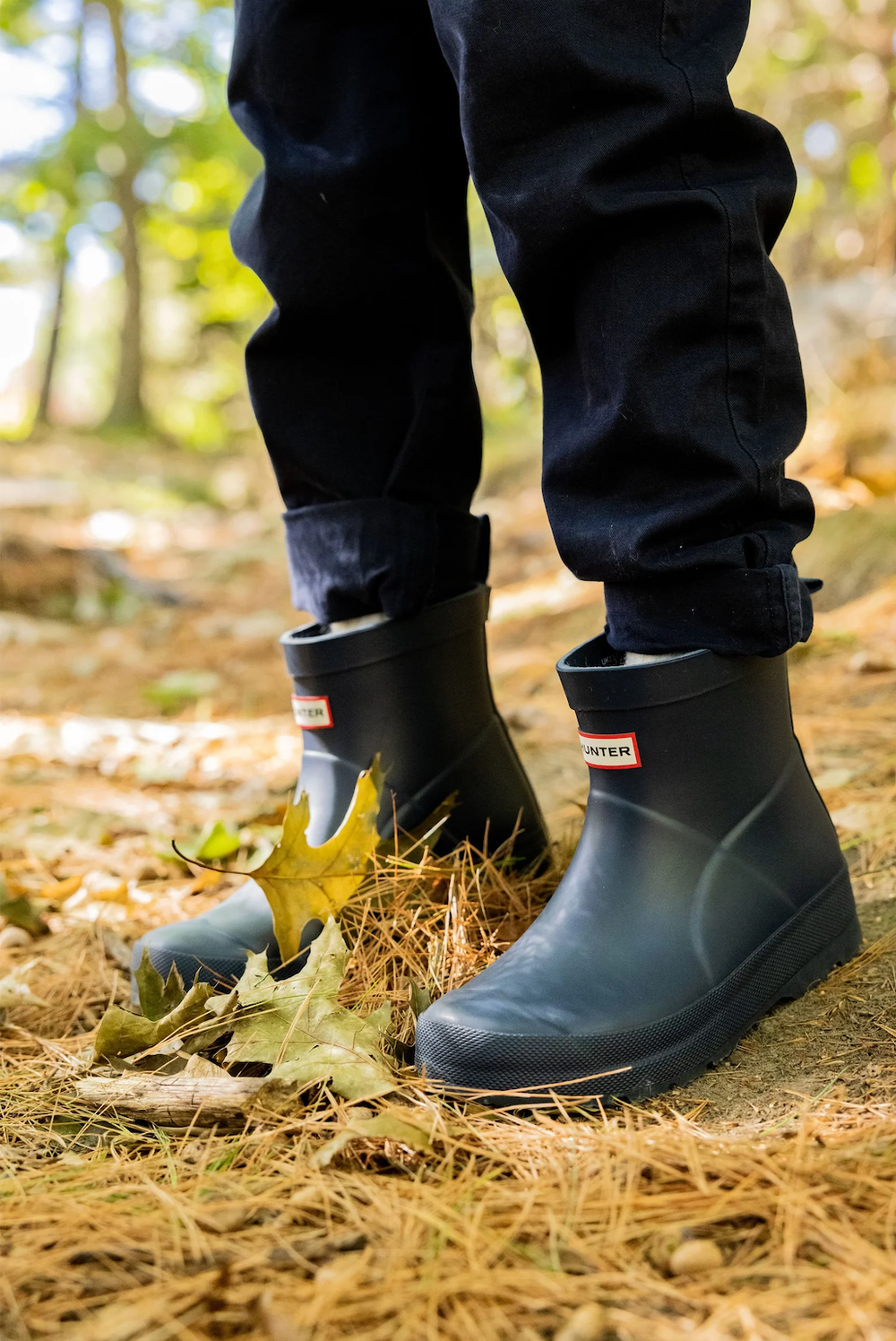 Close-up of a child wearing dark navy Hunter rain boots standing on a leaf-covered forest path.
