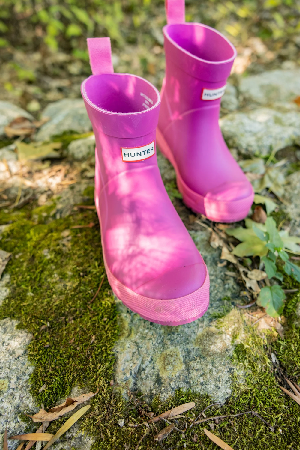 Pair of bright pink Hunter kids rain boots sitting on a mossy rock in a garden setting.