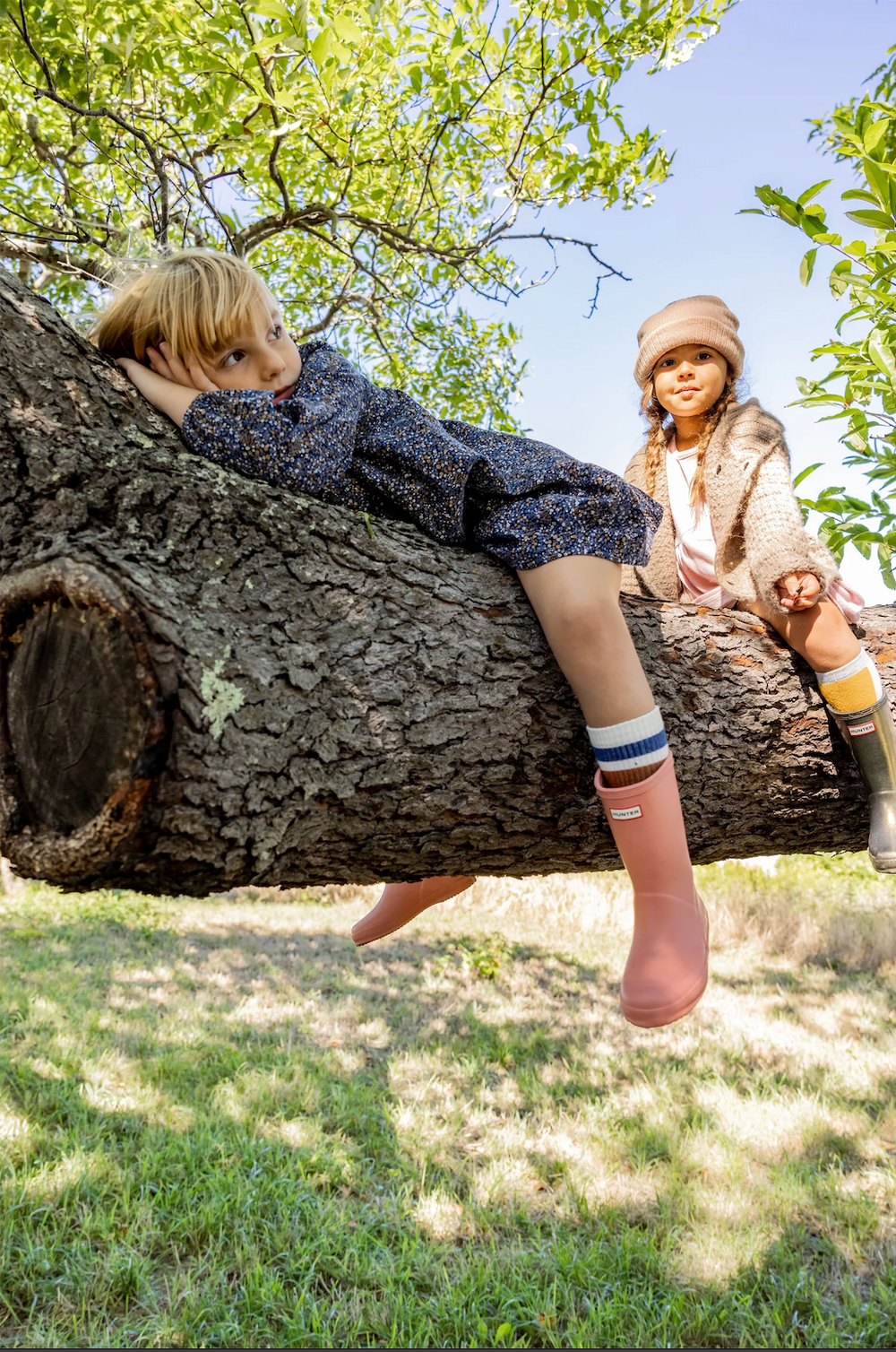 Two children climb a large tree trunk outdoors, one wearing pink striped Hunter rain boots.