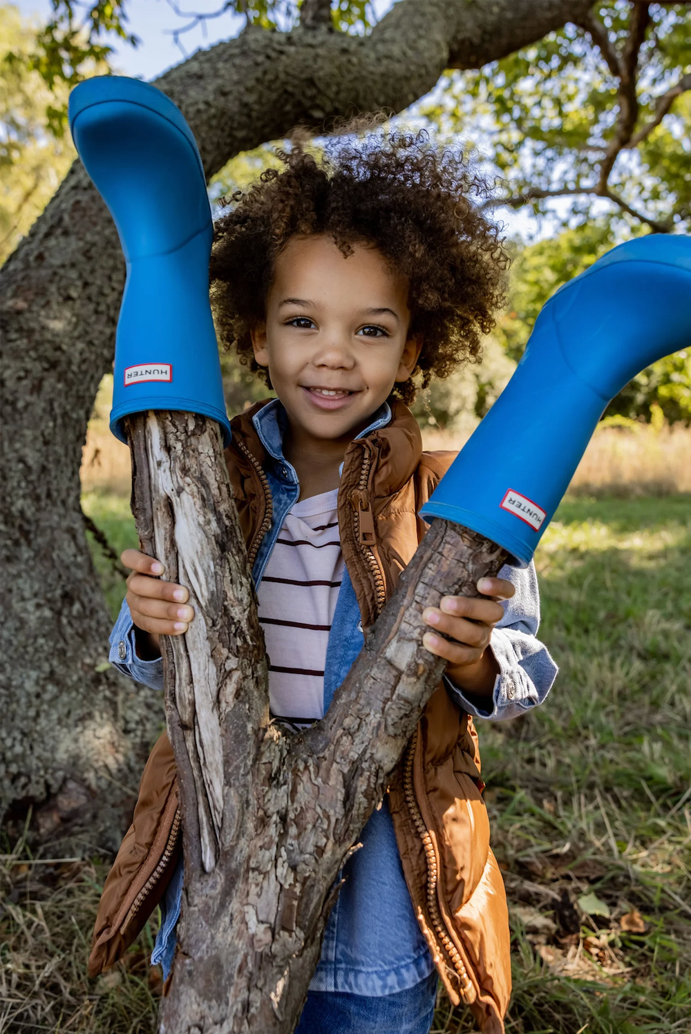 Smiling curly-haired child holds up blue Hunter rain boots while standing beside a tree outdoors.