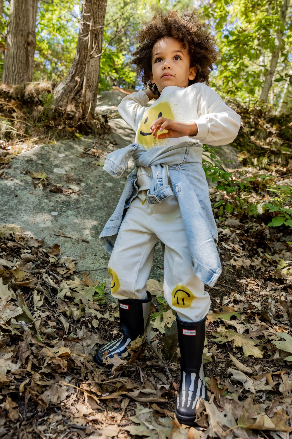 Young child in smiley face outfit and black Hunter rain boots exploring a forest trail covered in autumn leaves.