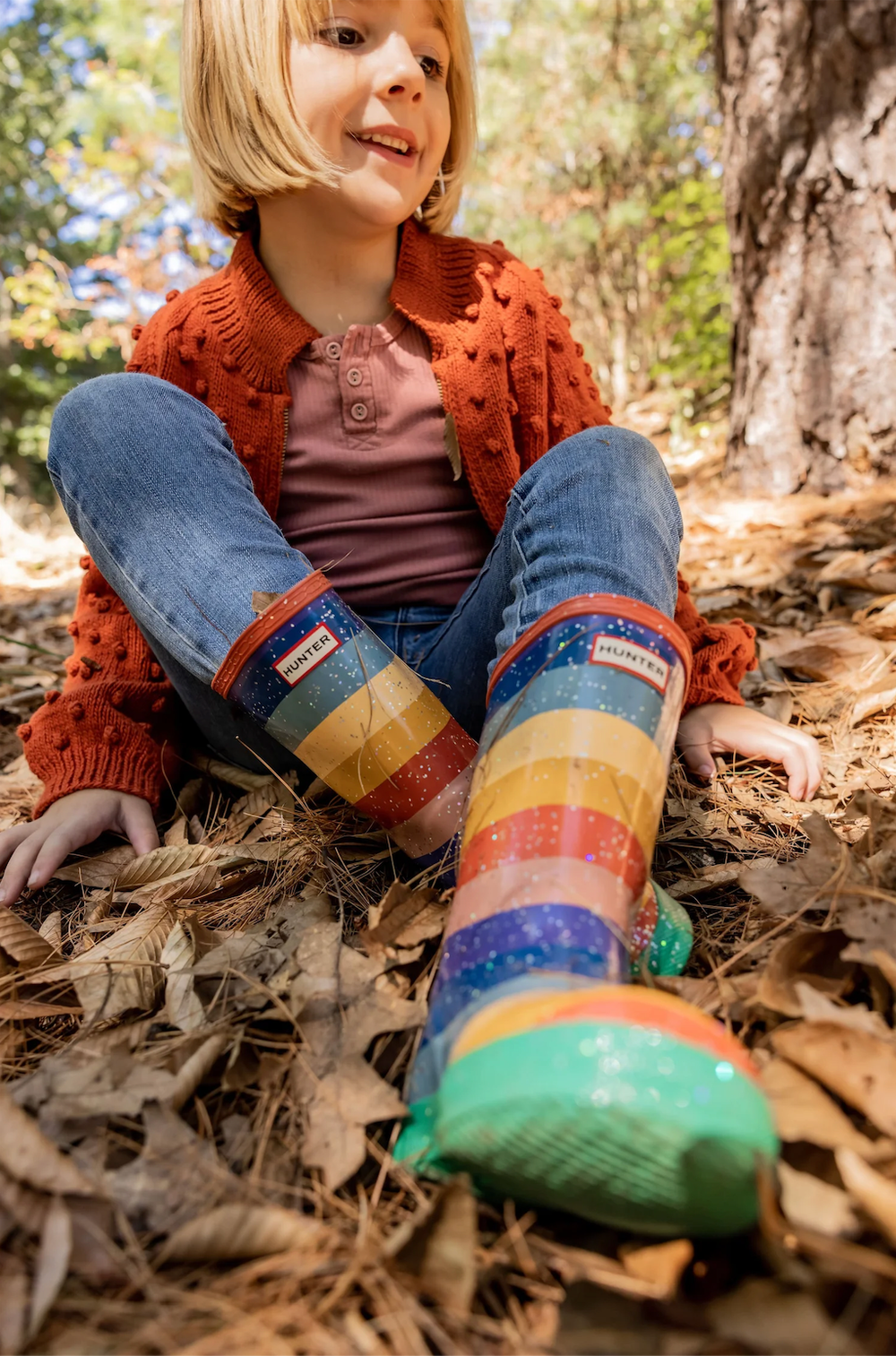 Smiling blonde child wearing rainbow striped Hunter rain boots sitting cross-legged on fallen autumn leaves.