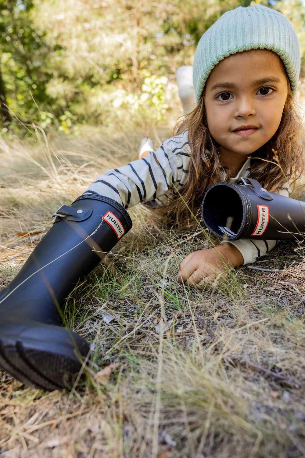 Young girl in black Hunter rain boots and striped shirt lying in dry grass in a sunlit forest.