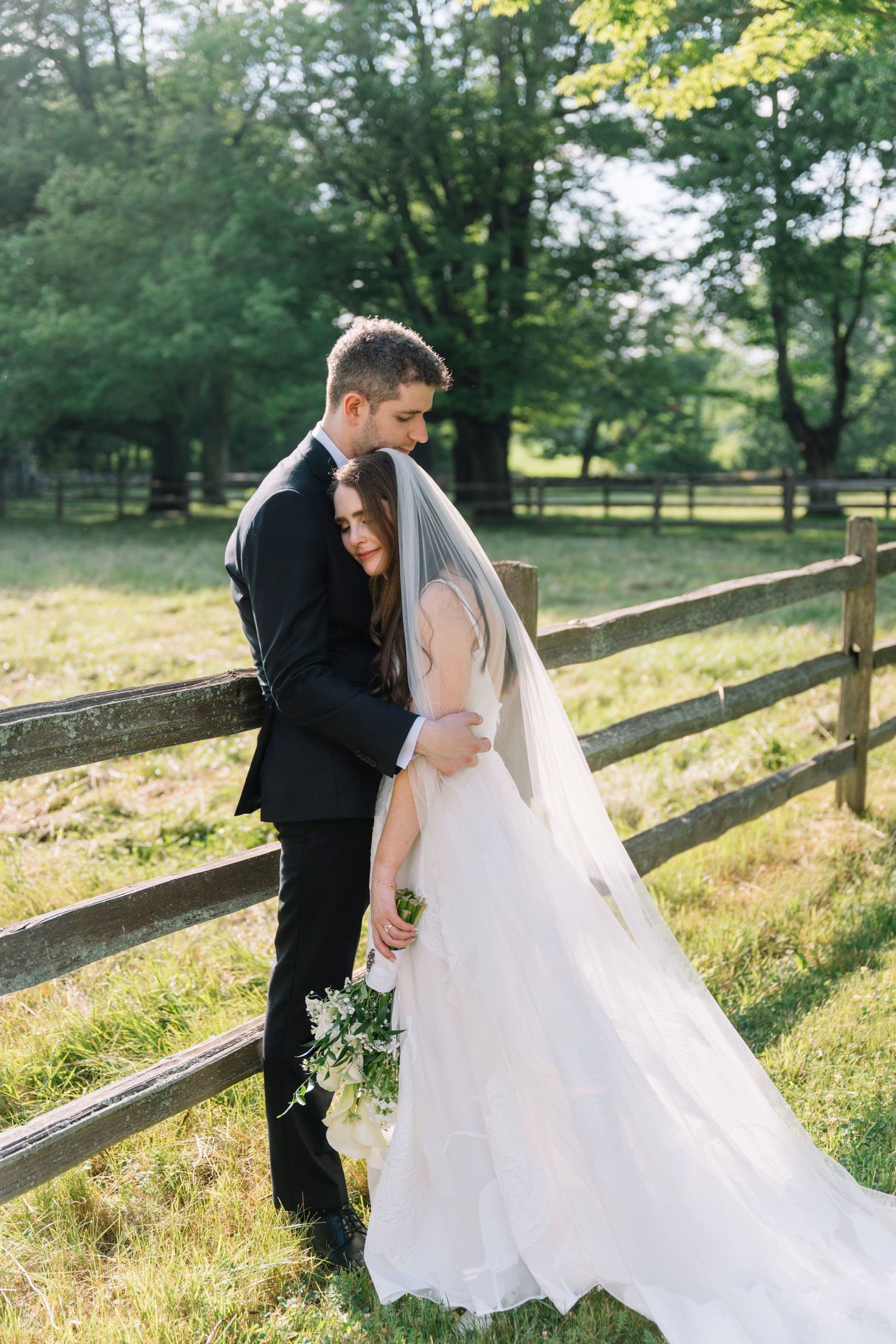 Bride and groom embrace by rustic wooden fence in lush green countryside, bride holding white bouquet in elegant wedding gown.