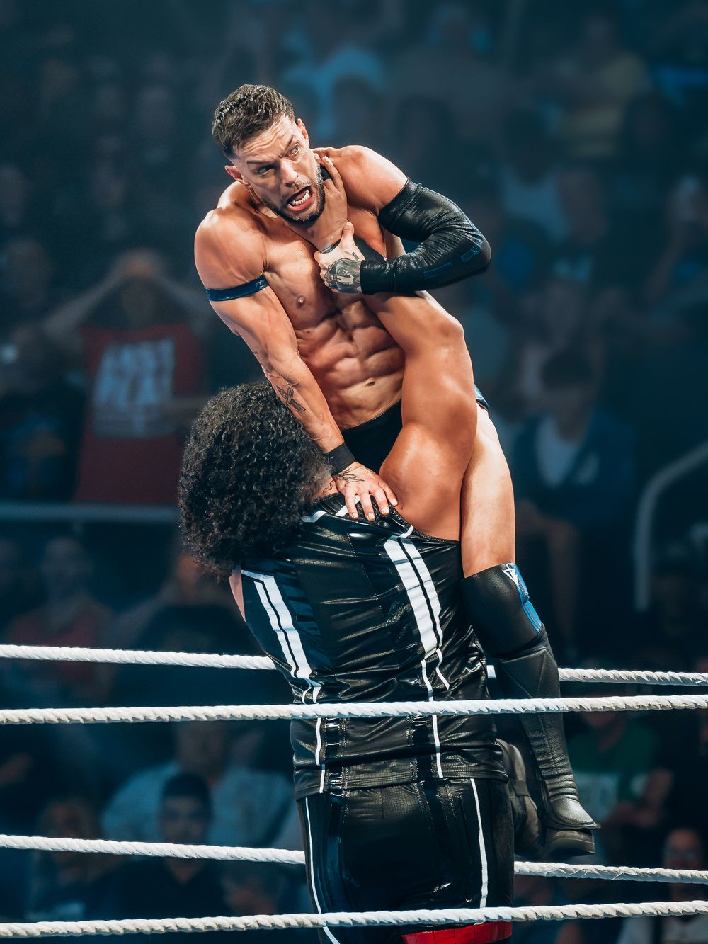 Two professional wrestlers grapple intensely in a WWE ring, one dominating from above while crowds watch in the background.