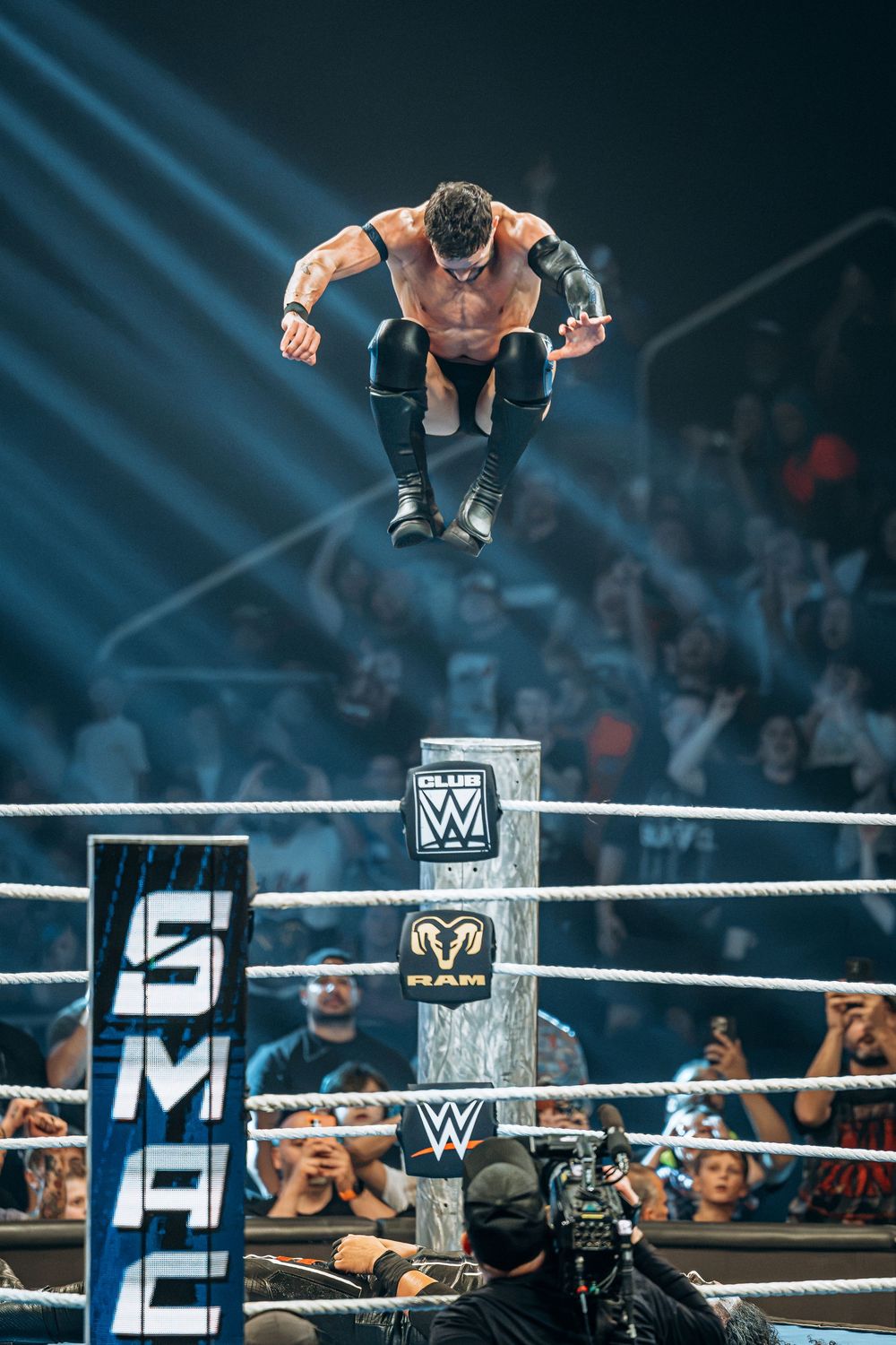 WWE wrestler leaps from turnbuckle at SmackDown live event, soaring above the ring ropes before a packed arena crowd.