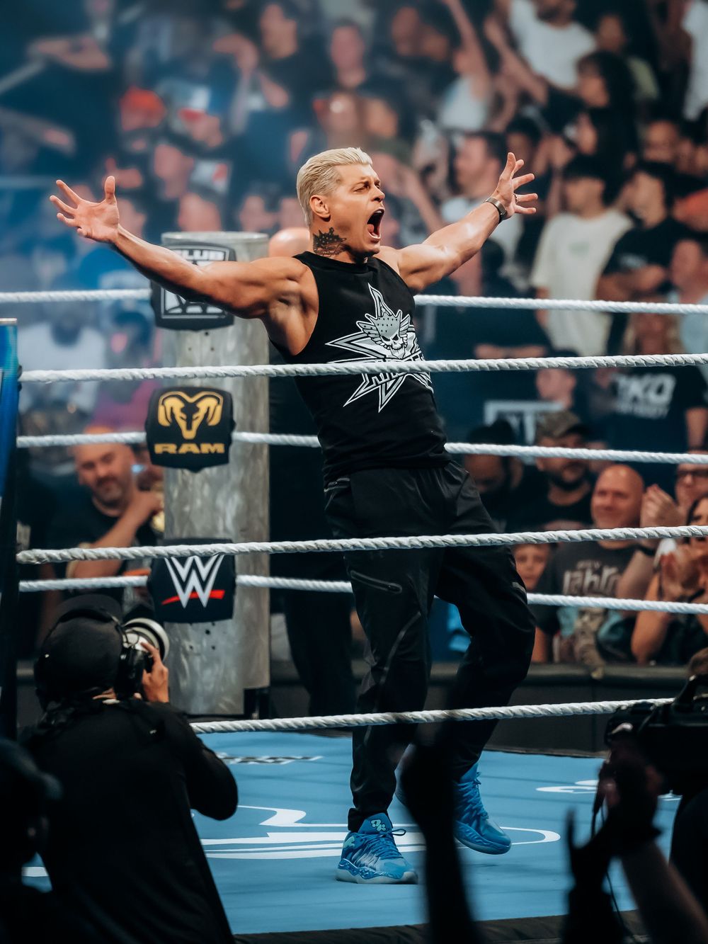 A WWE wrestler celebrates in the ring with arms raised, wearing a black tank top and blue sneakers, as the crowd cheers.