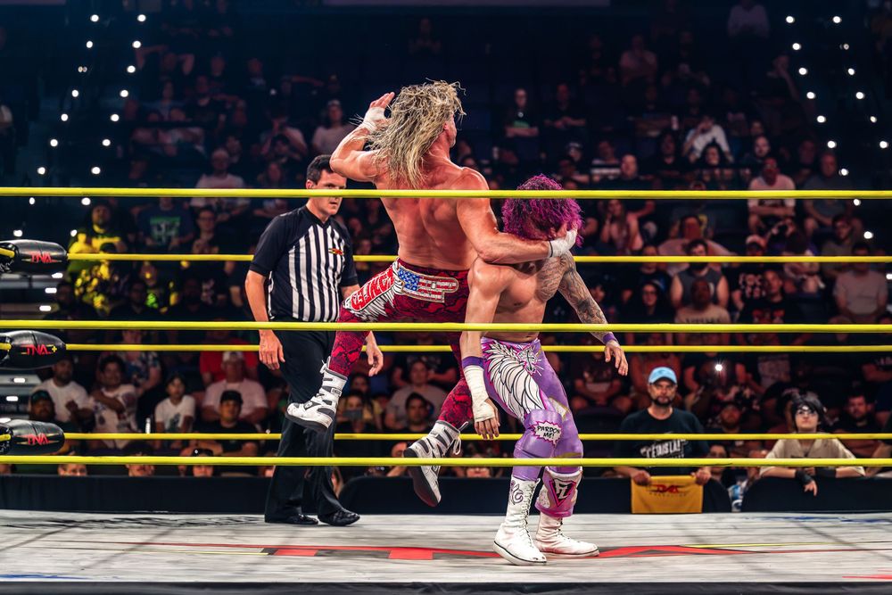 Two professional wrestlers grapple near yellow ring ropes as a referee watches during a WWE NXT event.