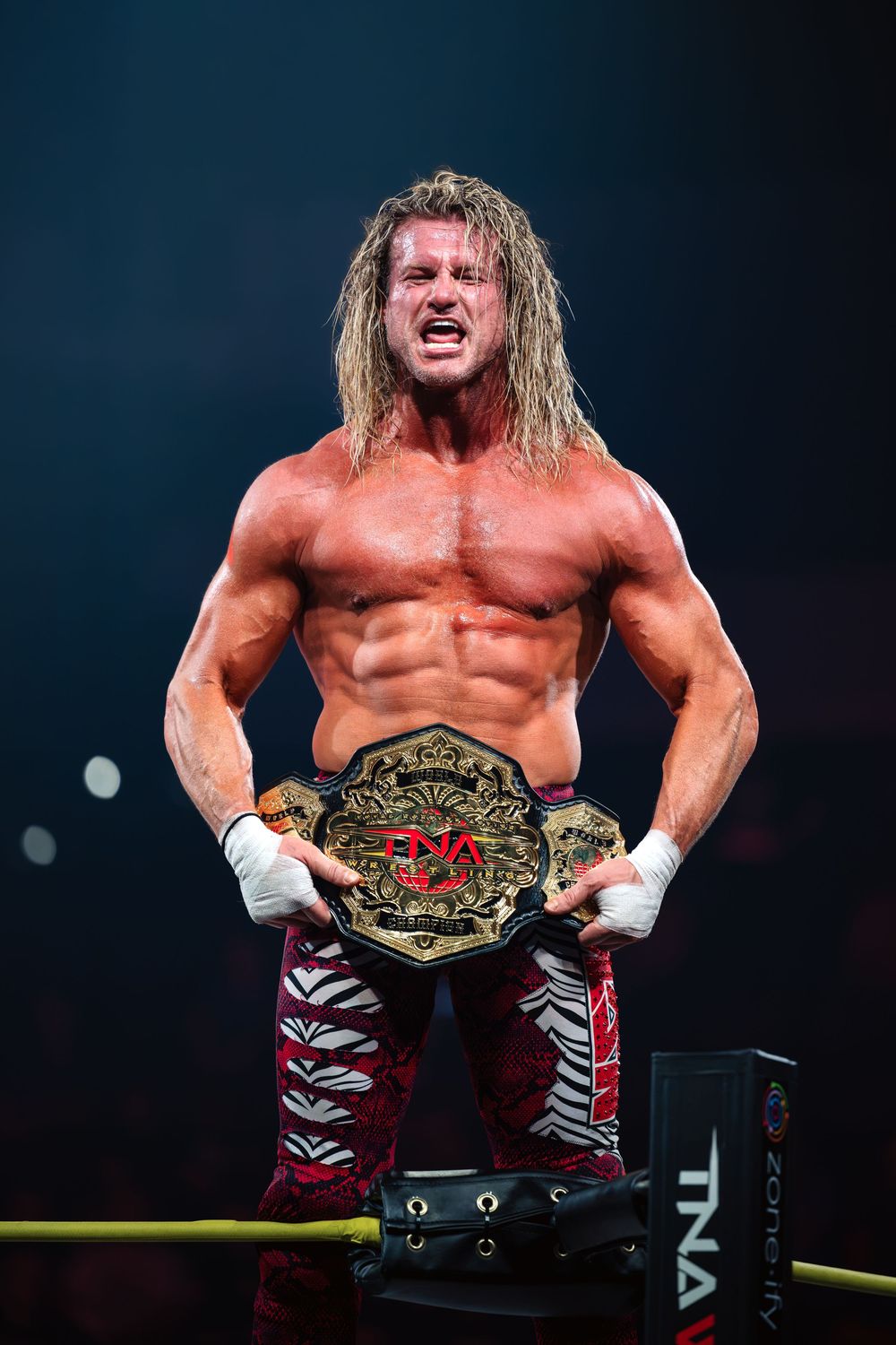 Muscular blonde wrestler holds TNA championship belt in wrestling ring.