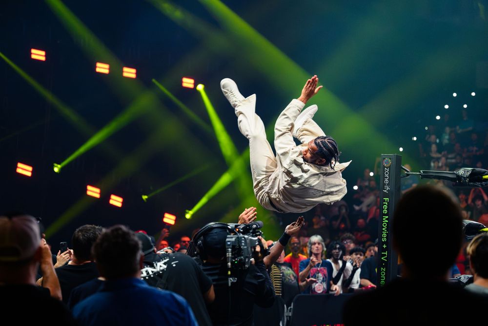 A performer executes an impressive aerial flip under vibrant green stage lights at a packed live wrestling event.