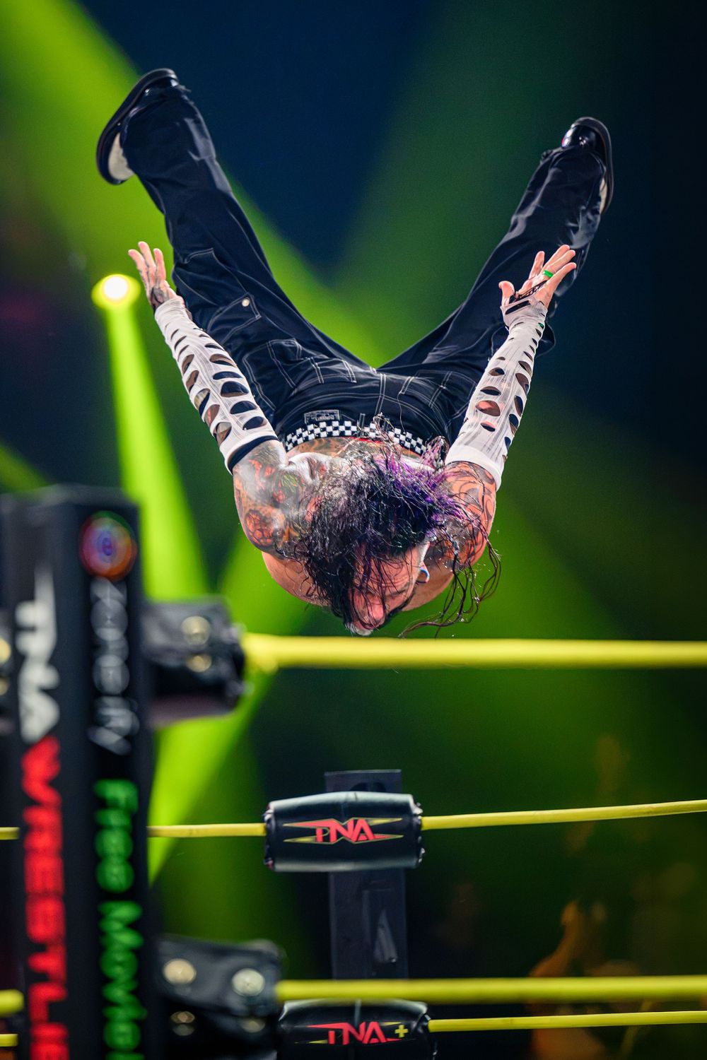 A tattooed TNA wrestler performs a dramatic dive over the ring ropes during a live wrestling event.
