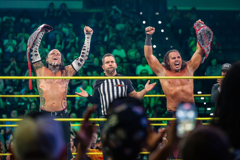 Two wrestlers celebrate a tag team championship victory, raising their belts under bright arena lights with a referee beside them.