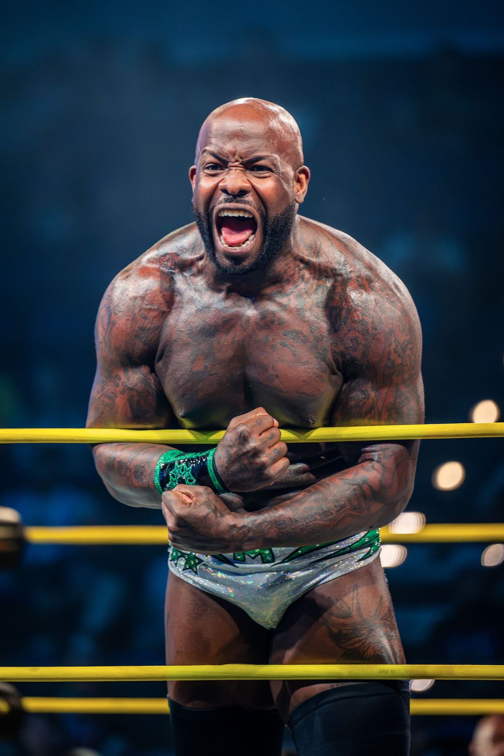 Muscular tattooed wrestler screams intensely while leaning on yellow ring ropes during a WWE NXT match.