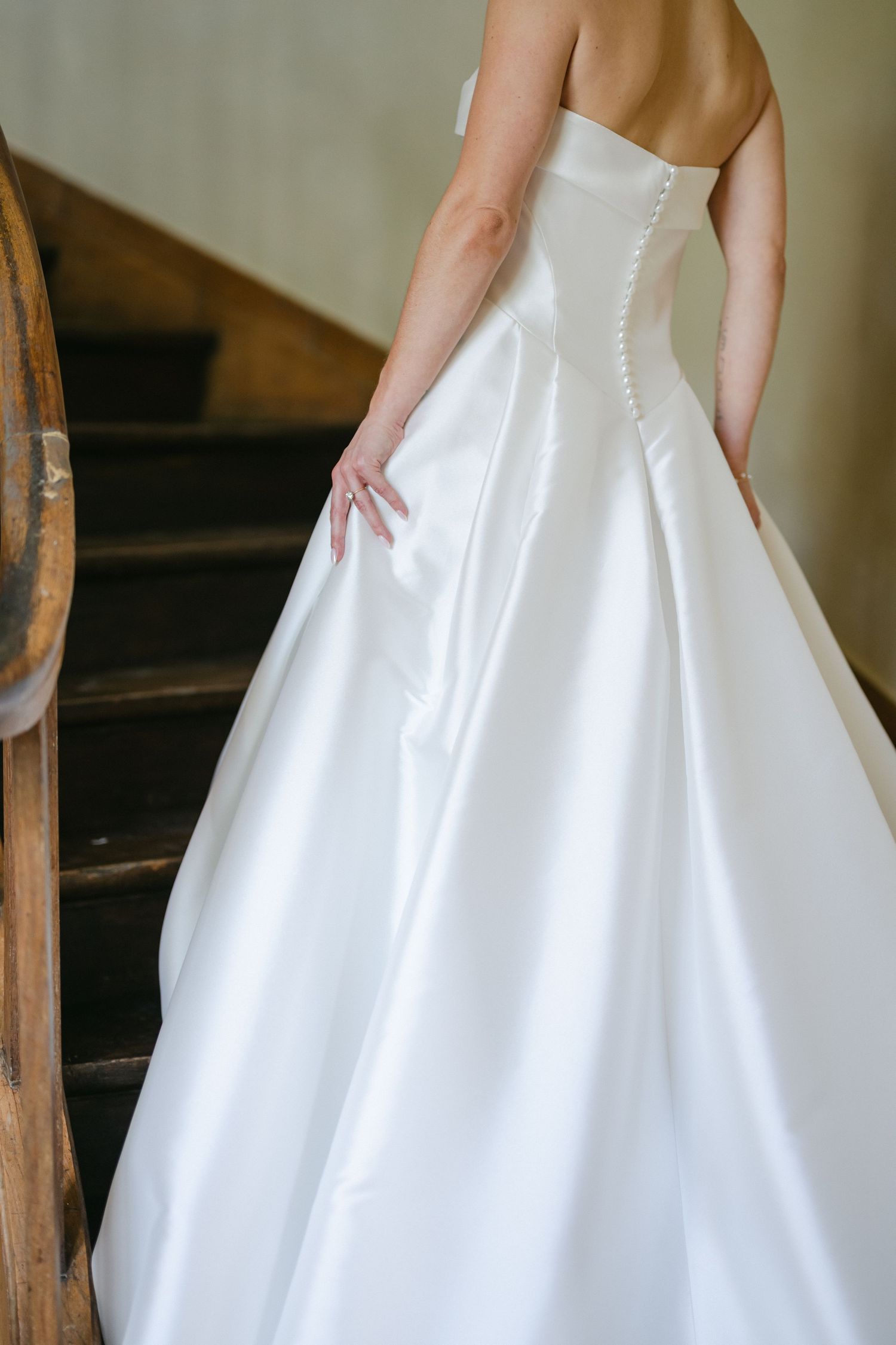 Bride in elegant strapless white satin ball gown with pearl button back detail, standing on wooden staircase.